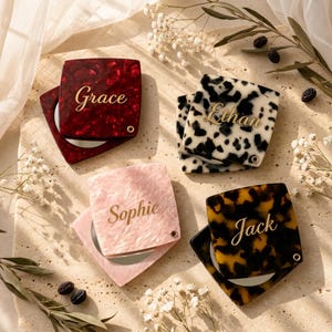 May include: Four square compact mirrors with hinged lids, each featuring a unique marbled pattern in red, black and white, light pink, and brown. Each lid is personalised with a gold-coloured name: Grace, Ethan, Sophie, and Jack.