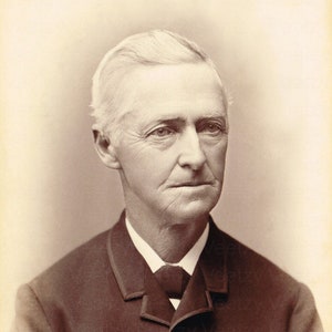 May include: A black and white portrait of an older man with white hair and a beard. He is wearing a dark suit jacket with a white shirt and a dark tie. The photo is in a vintage style.
