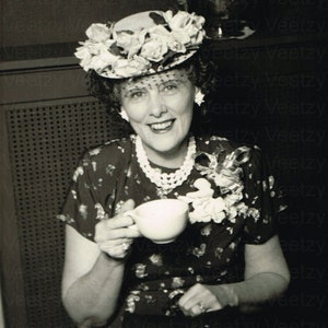May include: A woman wearing a floral hat and a patterned dress holds a white teacup. She is smiling and looking at the camera.