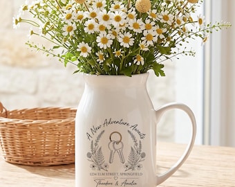 May include: A white ceramic pitcher vase filled with a bouquet of white daisies and yellow billy ball flowers. The pitcher features a black graphic of keys, a wreath, and the text A New Adventure Awaits, 1234 Elm Street, Springfield, Theodore & Amelia, 2026.