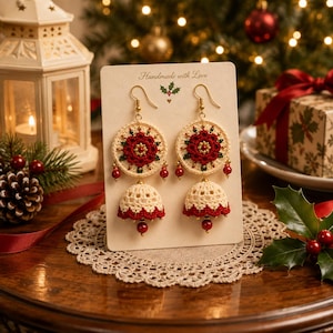 May include: A pair of cream and red crochet earrings featuring a circular top with a floral pattern and a bell-shaped bottom, accented with small red beads. The earrings are displayed on a rectangular card with the text Handmade with Love.