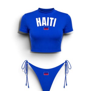 May include: A royal blue two-piece set consisting of a short-sleeved crop top and bikini bottoms. The top features the word HAITI in white block letters above a small Haitian flag. The bikini bottoms have side ties and a small Haitian flag emblem.