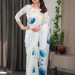 May include: A white saree featuring a blue floral print with large blue flowers and delicate leafy vines. The garment includes a matching long-sleeved blouse with a round neckline, showcasing a smooth fabric finish and a draped silhouette.