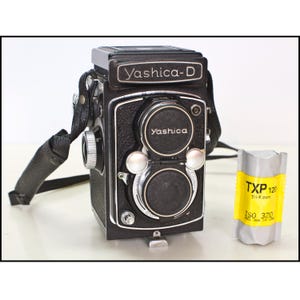 May include: A black Yashica-D twin-lens reflex camera with a leather strap. The camera has the name "Yashica-D" on top and "Yashica" on the lens covers. A roll of TXP 120 Tri-X pan film is next to the camera.