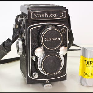 May include: A black Yashica-D twin-lens reflex camera with a black strap and a roll of TXP 120 Tri-X pan film. The camera has the name "Yashica-D" on the top and "Yashica" on the lens covers. The film is gray and yellow.