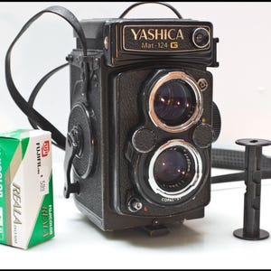 May include: A vintage Yashica Mat-124 G twin-lens reflex camera with a black leather strap, a roll of Fujicolor Reala 500 film, and a film spool. The camera has two lenses and the text "Yashica Mat-124 G".