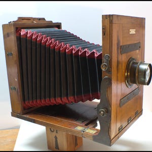 May include: Antique wooden camera with a bellows design. The camera features a lens, metal hardware, and a red and black bellows. The wooden body has a warm, natural finish. A classic photography item.