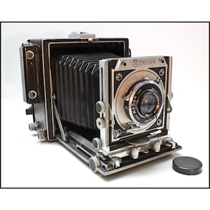 May include: A black and silver Meridian folding camera with a black accordion bellows, a Wollenak Rapax lens, and a Kalart rangefinder. The front plate features the Meridian logo and text, with various metal adjustment knobs and a separate black lens cap.