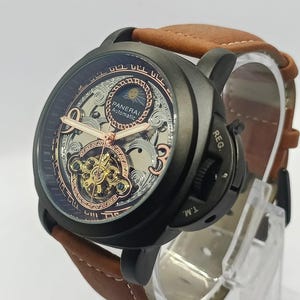 Może przedstawiać: Matowy czarny zegarek Panerai Automatic ze szkieletową tarczą, wskazówkami w kolorze różowego złota i wskaźnikiem faz księżyca. Zegarek posiada charakterystyczną osłonę koronki z napisem REG. T.M. oraz brązowy skórzany pasek z przeszyciami.