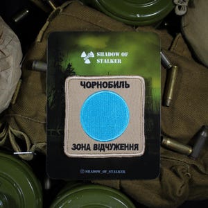 Op de afbeelding: Een vierkante geborduurde patch met een beige achtergrond en een felblauwe cirkel in het midden. De tekst "ЧОРНОБИЛЬ" is in het zwart boven de cirkel geborduurd en "ЗОНА ВІДЧУЖЕННЯ" is in het zwart onder de cirkel geborduurd.