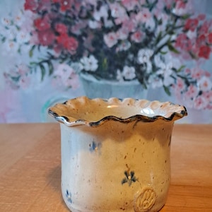 Könnte beinhalten: Ein kleiner, zylindrischer Keramikbecher mit einem gewellten, gerüschten Rand. Die Außenseite ist cremefarben gesprenkelt und mit kleinen, handgemalten blauen Blumenakzenten sowie einem kreisförmigen Prägestempel nahe der Basis versehen.