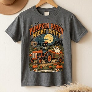 May include: A charcoal gray t-shirt featuring a graphic of a black cat driving an orange tractor in a pumpkin patch at night. The design includes a ghost, a full moon, and the text PUMPKIN PATCH NIGHT SHIFT and OPEN TILL THE LAST FULL MOON.