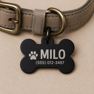 Puede incluir: Una placa de identificación para mascotas en forma de hueso negro mate unida a un collar de cuero color topo con herrajes de latón antiguo. La placa presenta un icono de huella grabado, el nombre MILO y el número (555) 012-3487 en letra sans-serif.