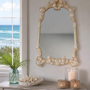 May include: A decorative mirror with a seashell design hangs above a wooden console table. The mirror has an arched shape and is adorned with various seashells. A glass vase with green leaves, a coral bowl, and candle holders complete the coastal decor.