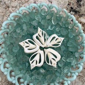 May include: Five white seashells arranged in a star shape on a bed of light blue sea glass in a decorative bowl.