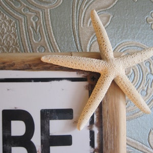 May include: A decorative wooden frame with a white sign and a starfish. The starfish is a light beige colour and is positioned on the top right corner of the frame. The sign has black letters on a white background.