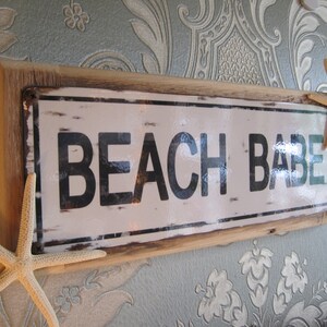 May include: A rectangular sign with the words "BEACH BABE" in black letters on a white background, framed by a light brown wooden border. Two starfish are attached to the sign.