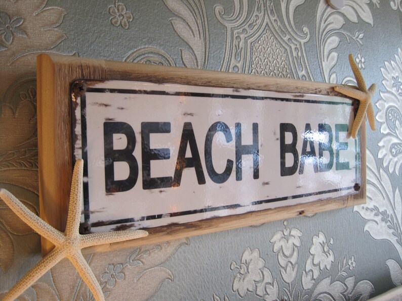 May include: A rectangular sign with the words "BEACH BABE" in black letters on a white background, framed by a light brown wooden border. Two starfish are attached to the sign.
