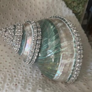 May include: A spiral seashell featuring a polished, iridescent surface with shades of teal, green, and white. The shell is decorated with rows of clear, sparkling rhinestones applied along the spiral ridges and the outer edge of the opening.