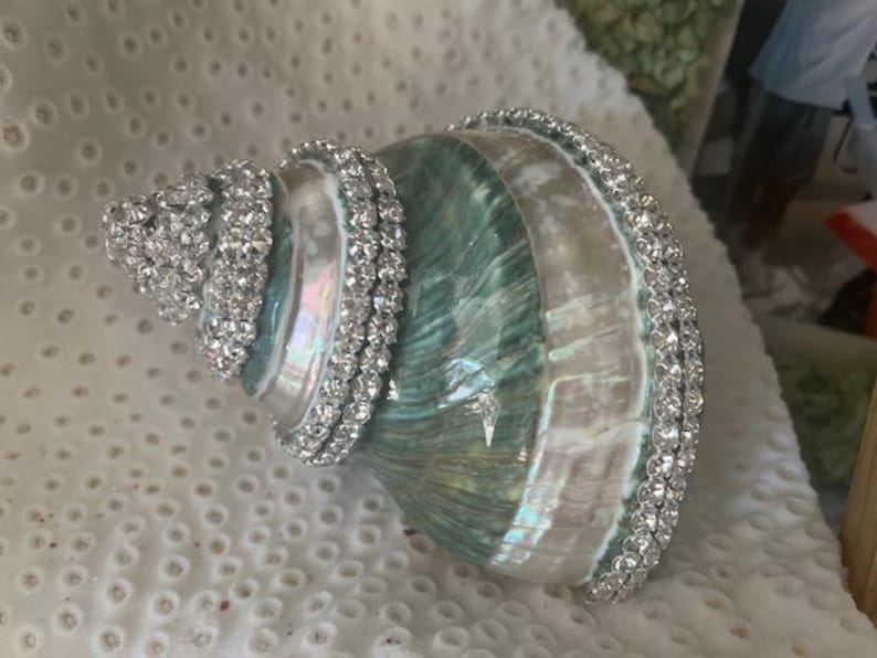 May include: A spiral seashell featuring a polished, iridescent surface with shades of teal, green, and white. The shell is decorated with rows of clear, sparkling rhinestones applied along the spiral ridges and the outer edge of the opening.