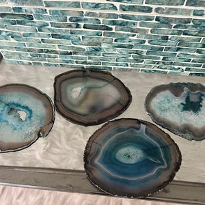 Puede incluir: Cuatro posavasos de ágata con un aspecto natural y pulido. Cada posavasos presenta un patrón único de tonos azules, turquesas y marrones, con un borde pulido. Los posavasos están dispuestos sobre una superficie de vidrio, con un fondo de mosaico de azulejos turquesa.