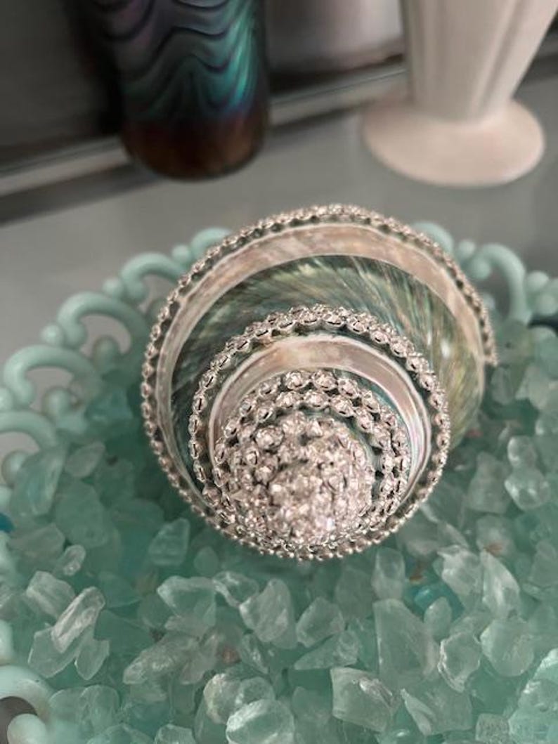May include: A spiral seashell decorated with rows of clear, sparkling rhinestones along its ridges. The shell features iridescent green and pearlescent tones, resting within a light blue decorative bowl filled with small, translucent blue glass stones.