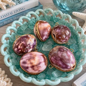May include: Four oval-shaped shell lockets with polished, mottled purple and white surfaces, each encased in a thin gold-toned metal frame with small loops at the top and bottom for attachment, resting inside a light blue scalloped glass bowl.