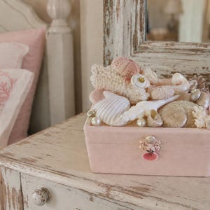 Pode incluir: Uma caixa de joias de veludo rosa cheia de conchas, corais e pérolas. A caixa tem um fecho decorativo e está sobre uma mesa de cabeceira branca desgastada. Uma garrafa decorativa e uma almofada estão no fundo.