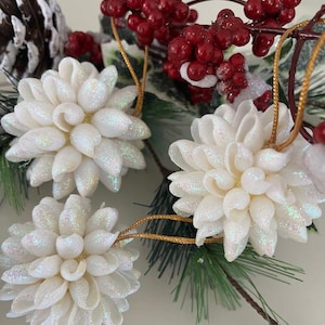 Puede incluir: Primer plano de adornos florales blancos cubiertos de purpurina con colgantes de cuerda dorada. Los adornos están rodeados de bayas rojas, agujas de pino y una piña, lo que sugiere un tema navideño.