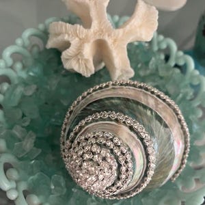 May include: A decorative seashell adorned with rows of clear rhinestones, resting in a light blue scalloped bowl filled with small light blue glass pebbles and accompanied by a white piece of coral placed behind the shell.