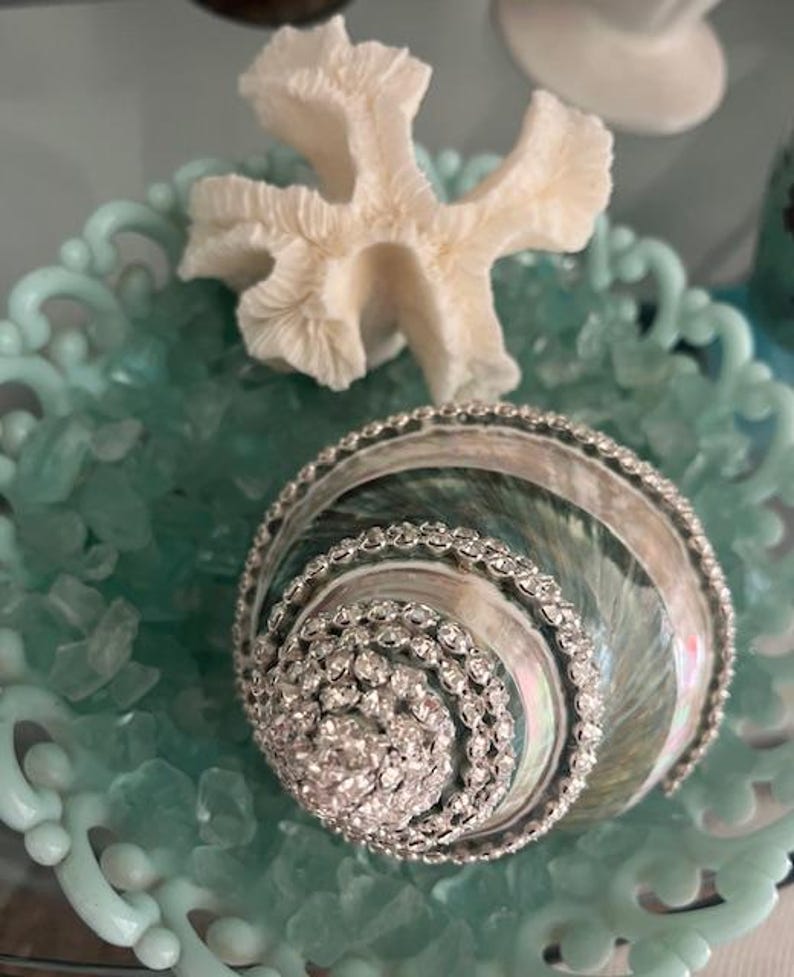 May include: A decorative seashell adorned with rows of clear rhinestones, resting in a light blue scalloped bowl filled with small light blue glass pebbles and accompanied by a white piece of coral placed behind the shell.