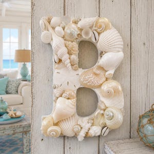 May include: A decorative letter B covered in a variety of white and cream-colored seashells, including spiral shells, bivalves, and a sand dollar. The shells are arranged to form the shape of the letter, creating a textured, multi-dimensional surface.