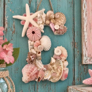 May include: A wall-hanging letter E decoration covered in various seashells, including a large white starfish at the top, pink sea urchin shells, and assorted spiral and bivalve shells in shades of white, cream, and soft pink, suspended by a rope.