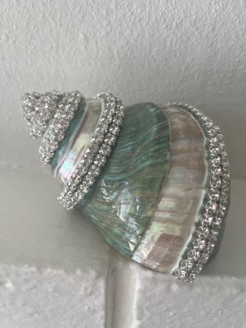 May include: A large spiral seashell featuring a natural iridescent finish with shades of teal, green, and cream. The shell is decorated with multiple rows of clear, sparkling rhinestones wrapped around the exterior ridges and the base edge.