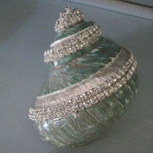 May include: A spiral seashell featuring a natural green and white textured surface, embellished with multiple rows of clear, sparkling rhinestones that follow the natural curves and ridges of the shell from the pointed apex to the wide base.