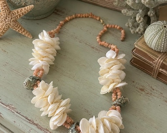 May include: A necklace featuring clusters of white, fan-shaped shells interspersed with small, light peach-colored beads. The design includes three larger, textured, greenish-grey spiral shells positioned at intervals along the strand.