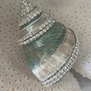 May include: A natural spiral seashell featuring an iridescent green and white surface, embellished with multiple rows of clear, sparkling rhinestones wrapped around the shell's exterior ridges and base, creating a decorative coastal accent piece.