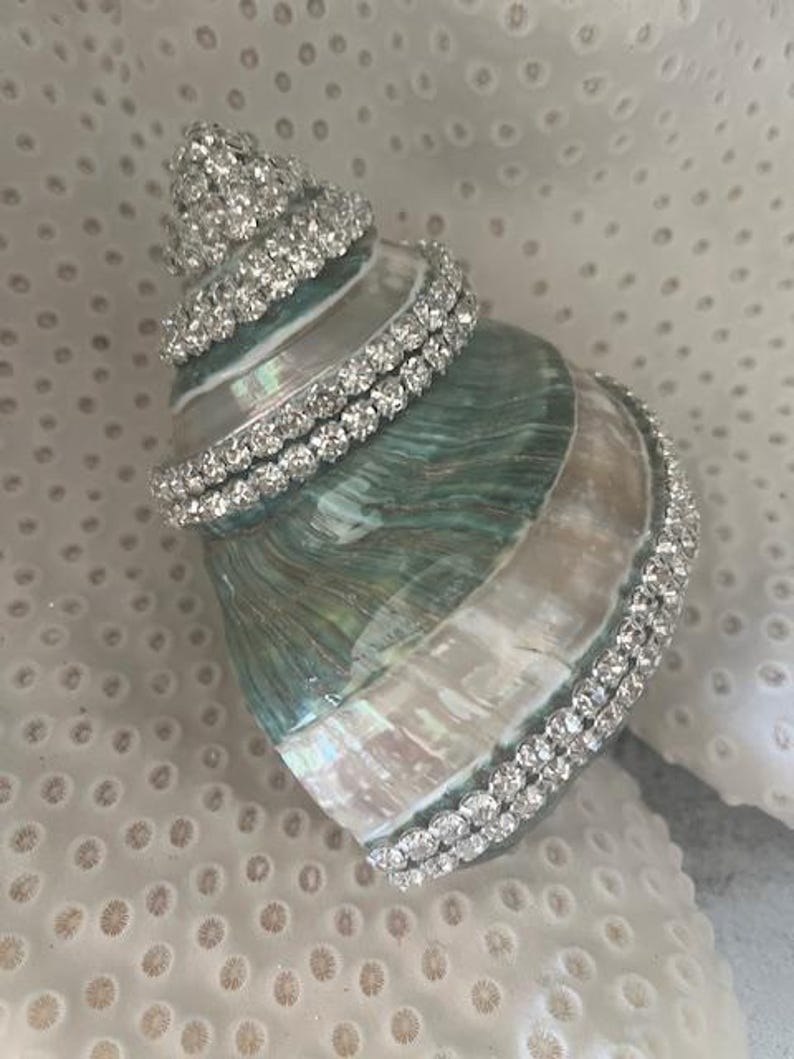 May include: A natural spiral seashell featuring an iridescent green and white surface, embellished with multiple rows of clear, sparkling rhinestones wrapped around the shell's exterior ridges and base, creating a decorative coastal accent piece.