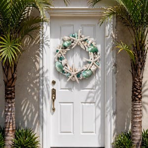 Op de afbeelding: Een witte voordeur met een decoratieve krans van schelpen en zeesterren. De krans heeft witte zeesterren, witte en turquoise schelpen en turquoise glazen kralen. Palmbomen omlijsten de deur, met groene planten aan de basis.