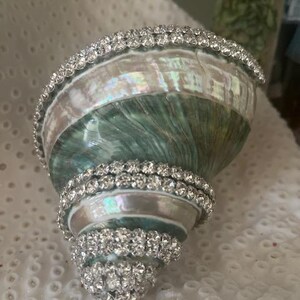 May include: A spiral seashell featuring an iridescent green and white pearlescent surface, decorated with multiple rows of clear, sparkling rhinestones wrapped around the shell's exterior ridges and along the wide opening's edge.