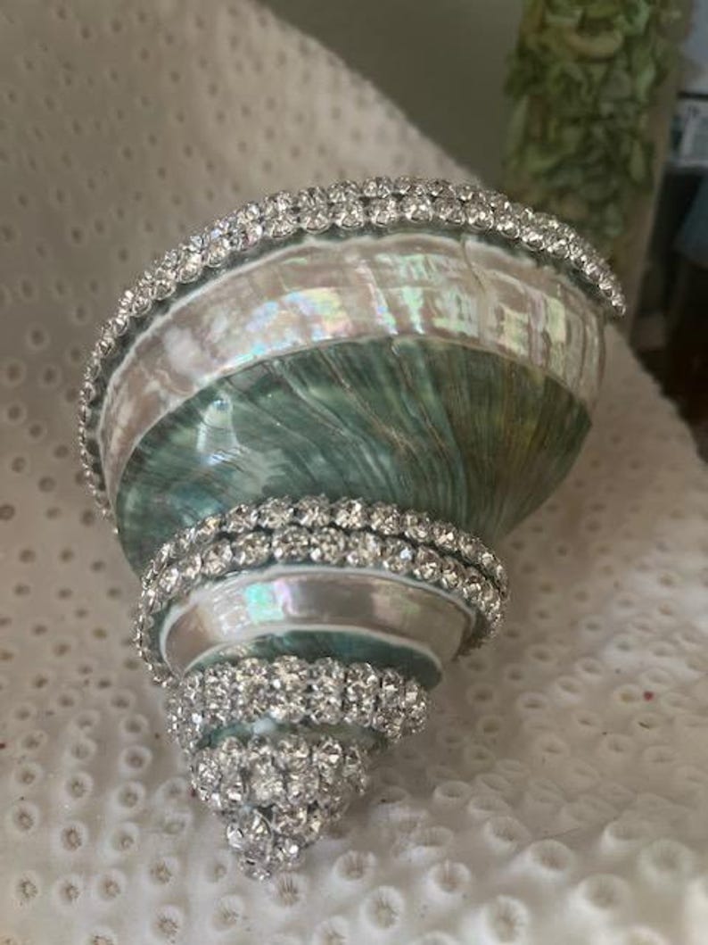 May include: A spiral seashell featuring an iridescent green and white pearlescent surface, decorated with multiple rows of clear, sparkling rhinestones wrapped around the shell's exterior ridges and along the wide opening's edge.