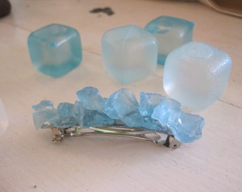 May include: A blue and clear glass hair clip with a silver bar. The clip is decorated with small, irregular pieces of glass that resemble ice shards.