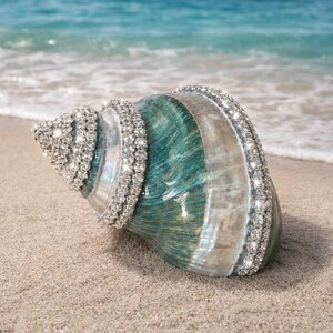 May include: A large spiral seashell featuring a polished teal and white gradient surface, decorated with multiple rows of sparkling clear rhinestones embedded along the natural ridges of the shell, creating a reflective and textured appearance.