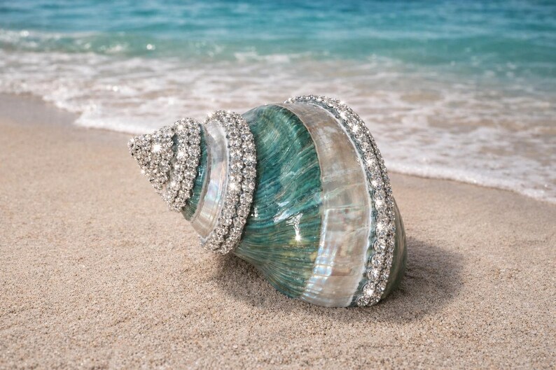 May include: A large spiral seashell featuring a polished teal and white gradient surface, decorated with multiple rows of sparkling clear rhinestones embedded along the natural ridges of the shell, creating a reflective and textured appearance.