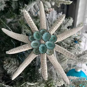 May include: A star-shaped ornament constructed from white, textured starfish arms arranged in a radial pattern. The center is adorned with a cluster of small, smooth, oval-shaped sea shells in shades of teal and blue with dark edges.