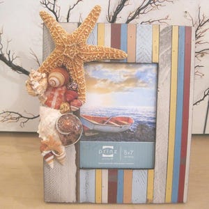 Pode incluir: Uma moldura de madeira branca com uma borda listrada multicolorida. A moldura tem um design de conchas e estrelas do mar e um inserto de foto de 13x18 cm. A foto mostra um barco na água.