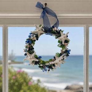 May include: A coastal-themed wreath with starfish, seashells, and blue accents. The wreath is adorned with white starfish, small seashells, and blue decorative elements. A blue ribbon hangs the wreath on a window, with a view of the ocean in the background.