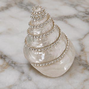 May include: A spiral-shaped seashell with a pearlescent white finish, adorned with a continuous strand of clear, sparkling rhinestones wrapped around the shell in a winding, upward-tapering pattern from the base to the pointed apex.