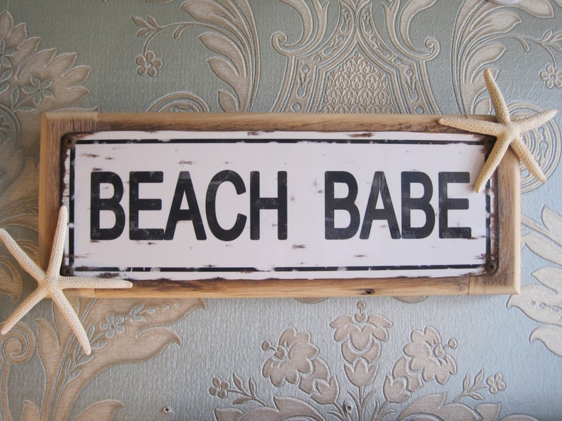 May include: A rectangular sign with the words "BEACH BABE" in bold black letters. The sign has a distressed white background and a wooden frame. Two starfish are placed on the sides of the sign, adding a coastal theme.
