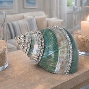 May include: A decorative seashell with a teal and white iridescent finish, embellished with rows of clear rhinestones. It is positioned between two clear glass cylindrical candle holders, each containing a white pillar candle, sand, and shells.